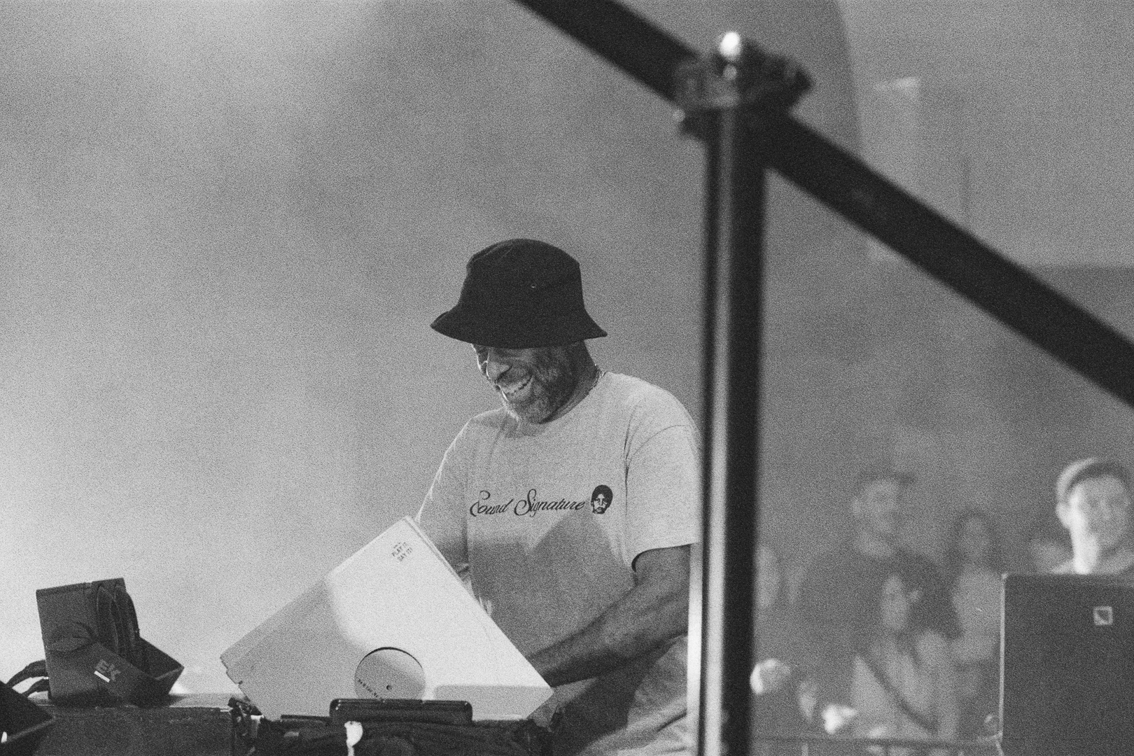 Black and white photo of Theo Parrish performing while smiling behind the booth. Beside him are a couple of records and in the background some audience members are present.